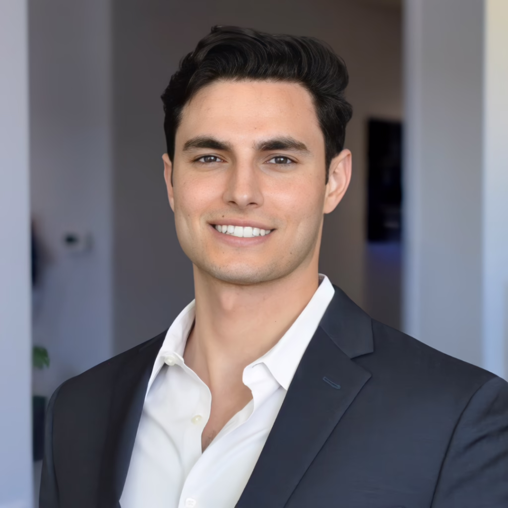 Professional headshot: man in a dark blazer and white shirt smiling at the camera in an office setting