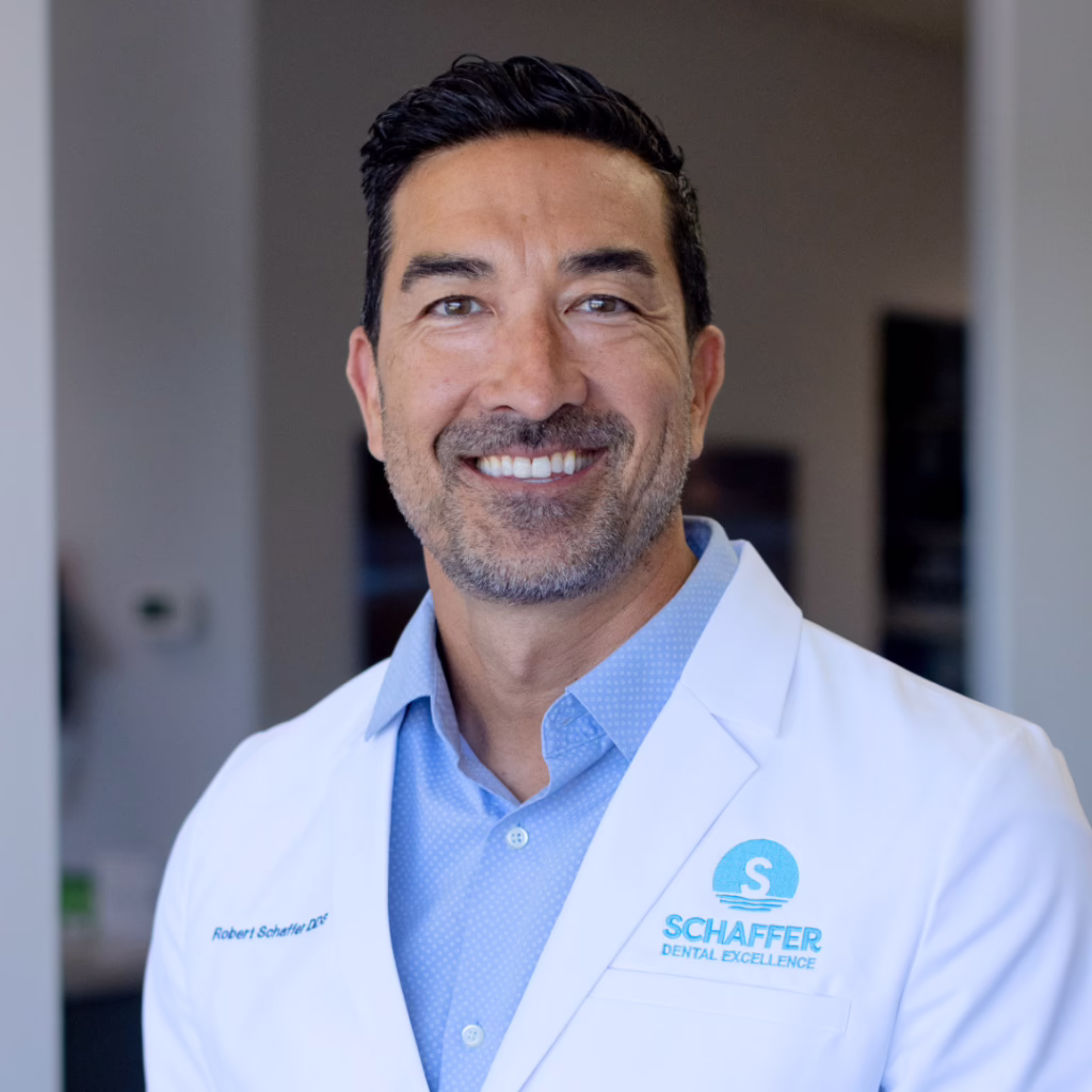Smiling male dentist in a white coat with Schaffer Dental Excellence logo, standing in a clinic background.