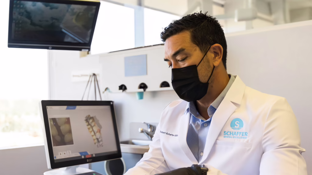 Dentist in a white coat and black mask examining dental X-ray images on a computer screen in a clinic.