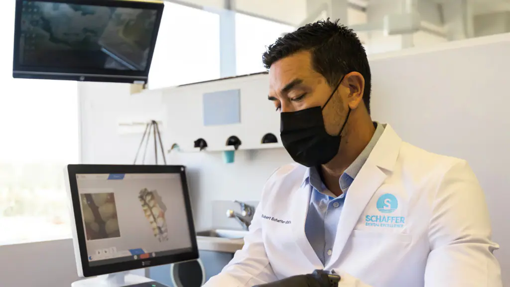 Dentist in a white coat and black mask examining dental X-ray images on a computer screen in a clinic.