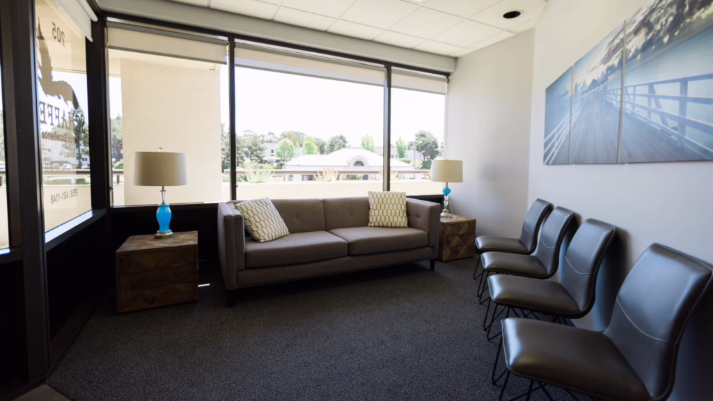 Modern office waiting area with a gray sofa, patterned pillows, wooden side tables with blue lamps, and a row of black chairs along the wall beneath a large window view outside.