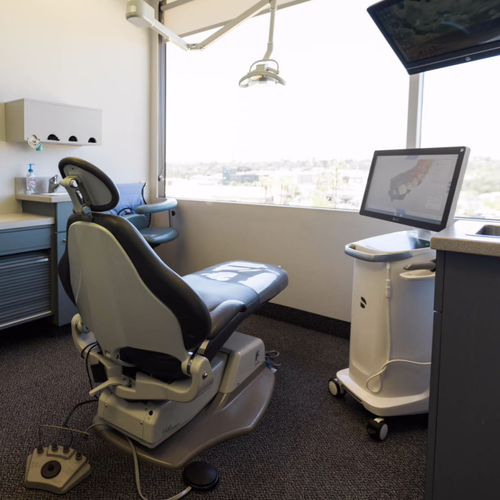 Dental exam chair in a clinic with a monitor on a mobile cart and overhead light, overlooking a window view to outside.