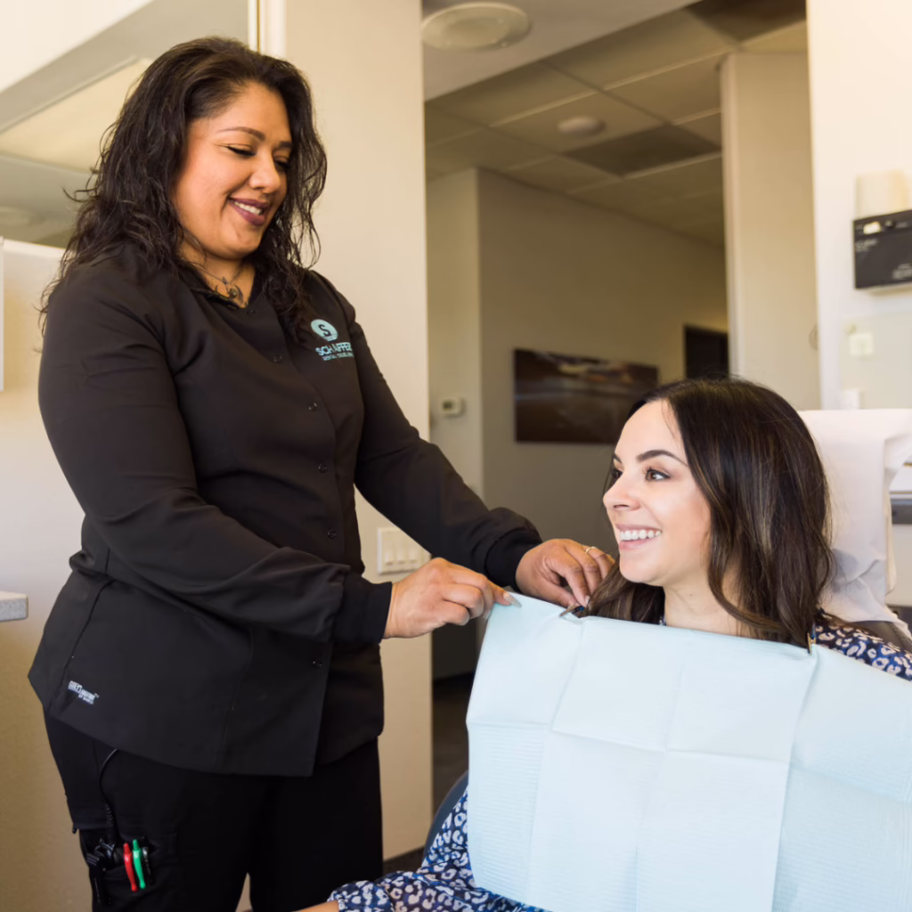 Dental assistant in a black uniform helps a smiling patient with a light blue bib in a clinic.