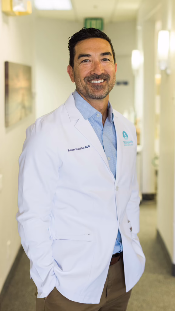 Smiling male dentist in a white coat standing in a bright clinic hallway, looking at the camera.